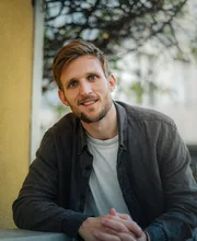 Alexander Hecking, founder coach, leaning forward on a ledge in the street and smiling